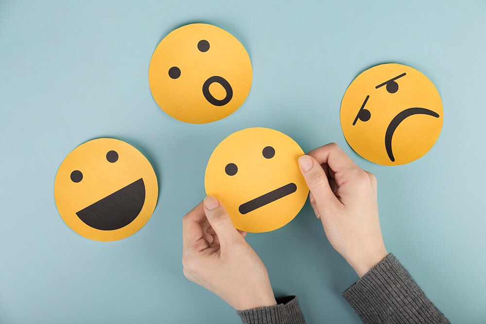Top view of hand arranging yellow paper emojis depicting happiness, awkwardness, sadness, and shock