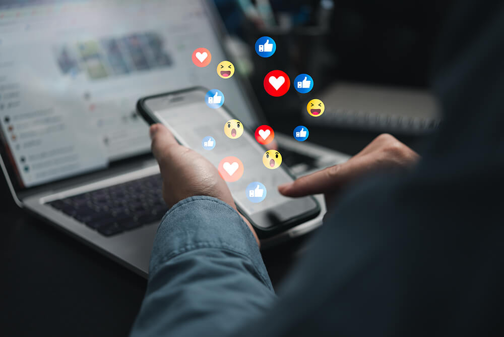 Person using a smartphone beside a laptop with floating social media reaction icons