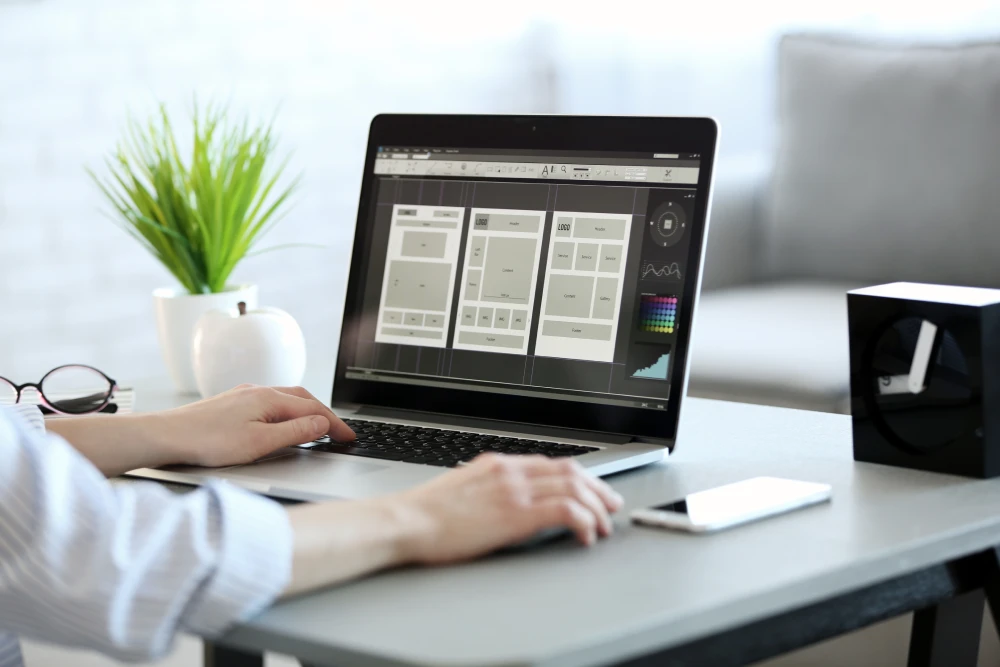 Close-up of a person working on a laptop displaying website UI wireframe layouts at a tidy desk.