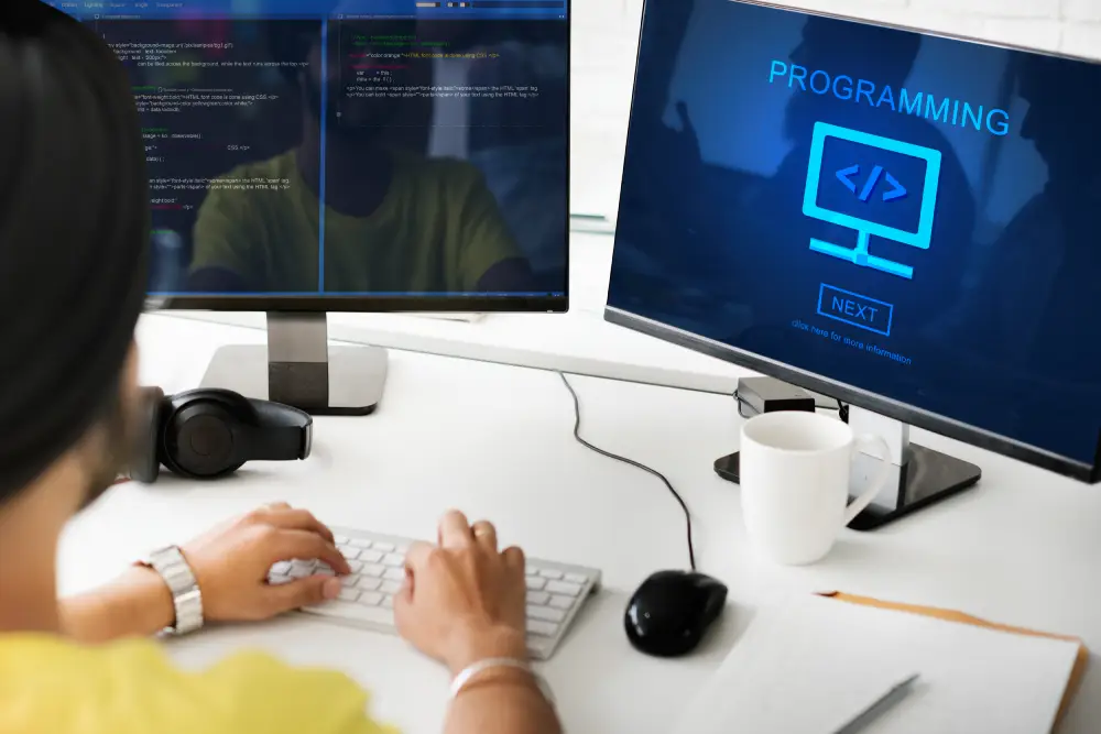 Developer typing on a keyboard at a dual-monitor workstation displaying code and a programming graphic.